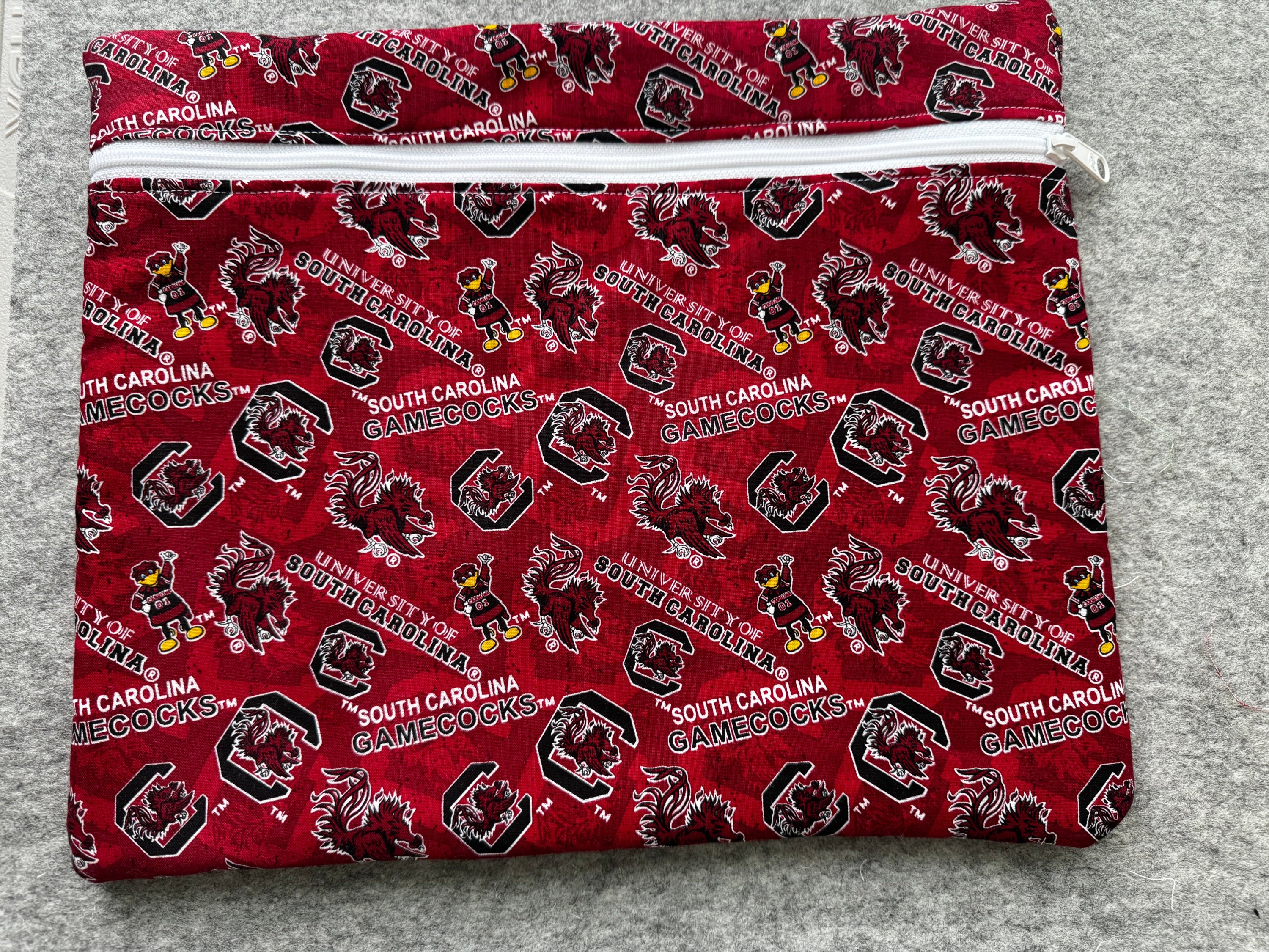 A folded fabric item, likely a pouch or sleeve, featuring a repeating South Carolina Gamecocks print in red, black, white, and yellow on a gray textured background.